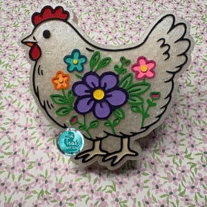 May include: A white chicken-shaped ornament with a red comb and wattle, featuring a colorful floral design with a large purple flower, a small orange flower, a teal flower, and a pink flower. A circular teal logo reads The Teal Mitten.