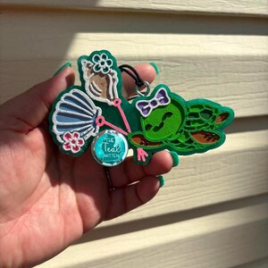 May include: A multi-layered 3D printed ornament featuring a green turtle with a purple bow, a blue seashell, a white conch shell with a flower, and a circular tag that reads The Teal Mitten. The design includes pink accents and a black hanging cord.