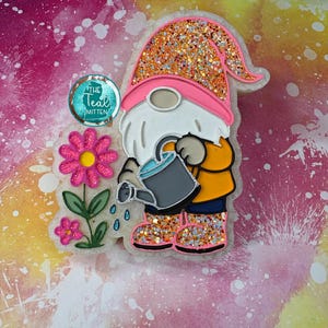 May include: A whimsical gnome figurine with a pink hat and glittery orange boots, watering a flower. The gnome has a white beard and is wearing an orange shirt and blue pants. A teal circle with the text "The Teal Mitten" is also present.