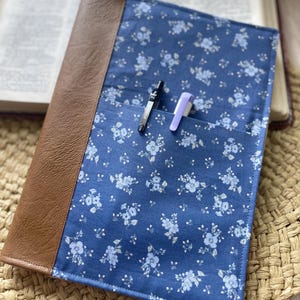 Può includere: Una copertina per libro rettangolare con un pannello in tessuto blu a motivo floreale bianco e un dorso in similpelle marrone. La parte anteriore presenta una tasca orizzontale contenente una penna nera e una penna viola chiaro.