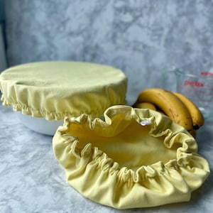May include: Two yellow fabric bowl covers with ruffled edges. One cover is on a white bowl, and the other is open, revealing the interior. A bunch of bananas and a measuring cup are in the background.