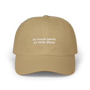 Könnte beinhalten: Beige Baseballcap mit gebogenem Schirm. Auf der Kappe steht in weißer Schrift "so much panic so little disco". Der Text ist in einer einfachen serifenlosen Schriftart gehalten. Die Kappe ist aus weichem Material.