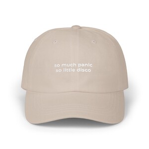 Könnte beinhalten: Beige Baseballkappe mit dem gestickten Text "so much panic so little disco" in Weiß. Die Kappe hat einen gebogenen Schirm und eine runde Krone, geeignet für den lässigen Gebrauch.