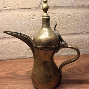 May include: A brass dallah coffee pot featuring a bulbous body, a long tapered spout, and a hinged lid topped with a decorative finial. The metal surface displays a weathered patina with visible signs of age and oxidation throughout the exterior.
