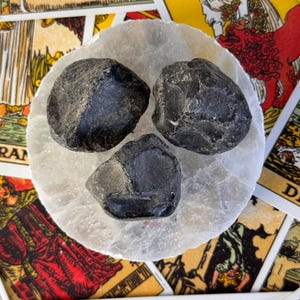 May include: Three dark gray, rough-textured stones rest on a white, circular, translucent surface. The stones are clustered together. Tarot cards with colorful illustrations and text are visible in the background.