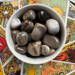 以下が含まれることがあります： グレー、ブラウン、クリーム色の滑らかで研磨された石が詰められた白いセラミックボウル。石にはユニークな模様と質感があります。ボウルはタロットカードで飾られた表面に置かれています。