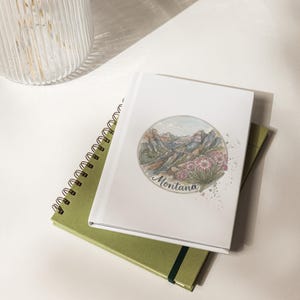 Puede incluir: Un cuaderno blanco con una ilustración en acuarela de un paisaje montañoso y la palabra "Montana". Está apilado sobre un cuaderno verde con espiral y un diario verde. Sobre una superficie blanca.