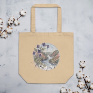 May include: Beige tote bag featuring a watercolor illustration of a river landscape with purple flowers and the text "New Jersey". The bag has a rectangular shape with a handle.