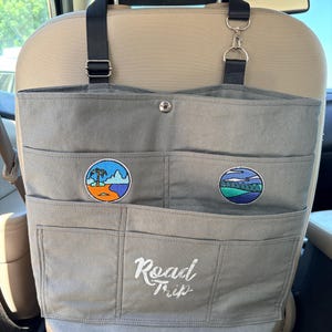 May include: A gray canvas car seat organizer featuring black adjustable straps with metal clips. The organizer has multiple pockets, two circular embroidered patches depicting a beach and a forest scene, and the white embroidered text Road Trip.
