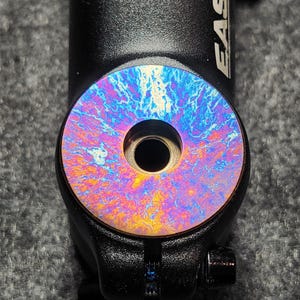 May include: Close-up of a black bicycle stem with a colorful, iridescent faceplate. The faceplate has a swirling pattern of blues, purples, and yellows. The word "EASTON" is visible on the side of the stem.