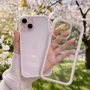 Könnte beinhalten: Eine transparente Handyhülle mit rosa Rand und einem rosa Telefon. Die Hülle wird vor einem Hintergrund aus weißen Blüten und grünen Blättern gehalten. Das Telefon hat zwei Kameralinsen.