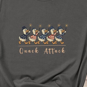 May include: Dark gray t-shirt featuring a whimsical design of marching ducks in patriotic attire, each holding a sparkler. The phrase "Quack Attack" is printed below the ducks in a playful font. The shirt is made of a soft material.