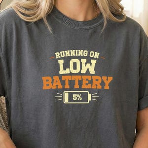 May include: Pepper-colored t-shirt with the text "RUNNING ON LOW BATTERY" in orange and yellow. The design includes a battery icon showing 5% charge. The shirt has rolled-up sleeves.