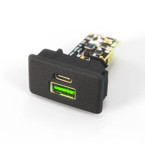 May include: A black rectangular panel-mount charging module featuring one USB-C port and one bright green USB-A port. The front face is textured black plastic, while the rear reveals an exposed circuit board with various electronic components.