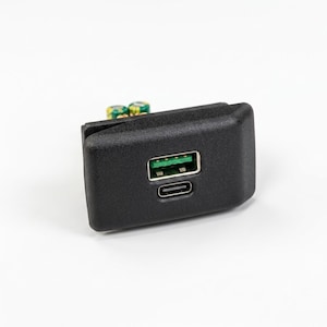Könnte beinhalten: Ein schwarzes, rechteckiges Elektronikmodul mit einem Standard-USB-A-Anschluss und einem kleineren USB-C-Anschluss auf der Vorderseite. Die Komponente hat eine matte Oberfläche und zeigt rückseitig grüne Leiterplatten und kleine Kondensatoren.