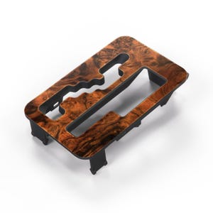 Puede incluir: Un embellecedor interior de coche con un patrón de vetas de madera. El embellecedor es rectangular con secciones recortadas y un borde negro. La veta de la madera presenta tonos marrones y negros, creando una apariencia natural y texturizada. La pieza está diseñada para encajar alrededor de una palanca de cambios.