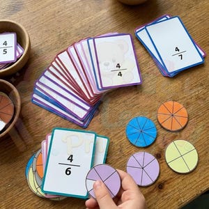May include: Educational fraction learning set with cards and circular fraction pieces. The cards display fractions like 4/4, 4/5, 4/6, and 4/7. The circular pieces are divided into segments and colored in blue, orange, purple, and yellow.