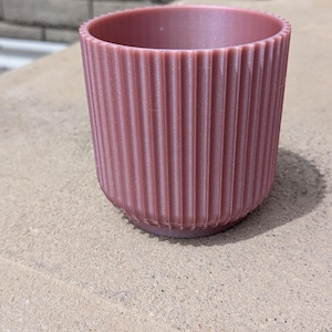 May include: A mauve-colored, cylindrical planter with vertical ribbed detailing. The small, round pot is made of a solid material and sits on a light-colored surface. The planter has a simple, modern design.
