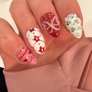 May include: Close-up of a hand with almond-shaped nails, each painted with a unique design. The nails feature red and white floral patterns, hearts, and a bow design. The overall color scheme is red, white, and pink.