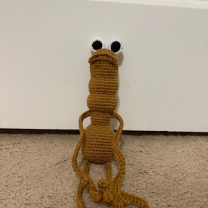 May include: A handmade, crochet creature in shades of brown. The whimsical figure has large, white-rimmed eyes with black pupils, a long neck, and outstretched arms and legs. The toy is made of yarn and sits on a neutral-colored carpet.