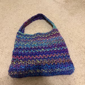 May include: A handmade crochet handbag featuring a variegated yarn pattern in shades of deep purple, bright blue, and neon yellow. The bag has a single integrated shoulder strap and a rectangular body with a textured, repeating stitch pattern throughout.