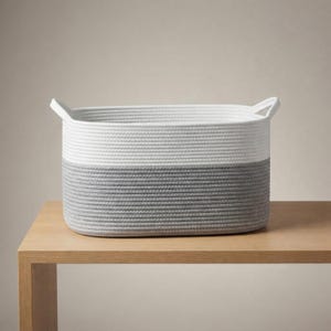 May include: An oval storage basket constructed from thick, coiled rope. The upper half of the basket is white, while the lower half is a medium gray. It features two integrated handles on the sides, forming a continuous, textured, and durable design.