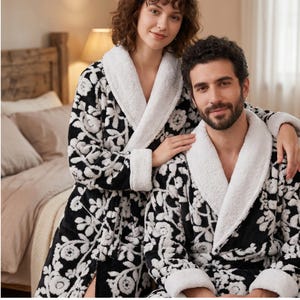 May include: Two plush bathrobes featuring a black base with a white floral pattern, long sleeves, and a thick white sherpa-style shawl collar. The robes are made of a soft, textured fabric and include a matching tie belt at the waist for closure.