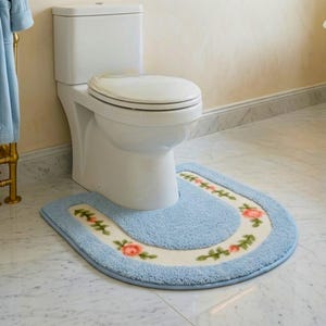 Könnte beinhalten: Hellblauer, U-förmiger Badezimmerteppich mit floralem Design. Der Teppich hat einen hellblauen Rand und eine cremefarbene Mitte mit rosa Rosen und grünen Blättern. Der Teppich liegt vor einer weißen Toilette.