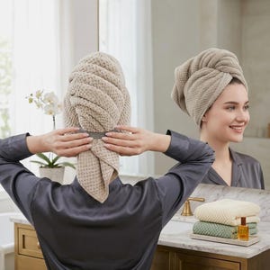Pode incluir: Uma pessoa está usando uma toalha de cabelo bege com tecido waffle. A pessoa está vestindo um roupão de cetim cinza. Um espelho reflete a pessoa e uma pilha de toalhas bege e verdes.