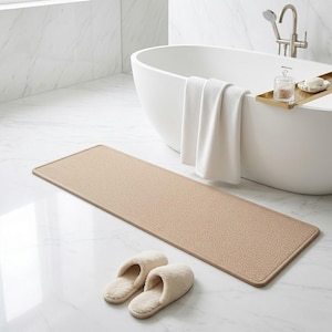 May include: A beige rectangular bath mat with a textured surface on a marble floor. Two cream-coloured, fluffy slippers sit near the mat. A white bathtub and a folded white towel are in the background.