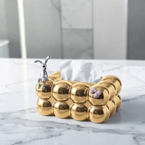 May include: A rectangular tissue box holder with a metallic gold finish and a bubble-like texture. A small, silver-colored rabbit figurine sits on the top left corner, and a light pink bow is attached to the right side. White tissues are visible.