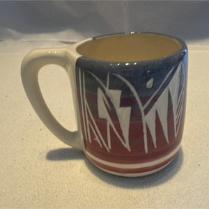 May include: A ceramic mug with a cream-colored handle and interior. The exterior features a design with red, white, and blue geometric shapes. The mug has a cylindrical shape and is suitable for hot or cold beverages.