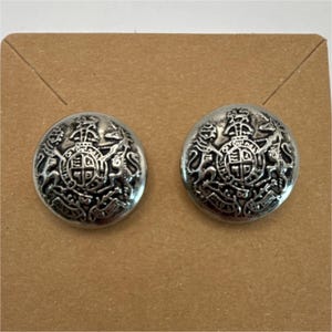May include: Two circular silver-toned metal buttons featuring a raised relief design of a royal coat of arms. The emblem depicts a central shield flanked by a lion and a unicorn, with intricate detailing and a polished metallic finish throughout.