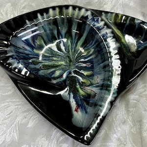May include: A black ceramic ashtray with a unique, swirling design. The interior features a blend of blue, green, and white colours, creating a marbled effect. The ashtray has a distinctive, curved shape with a glossy finish.