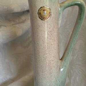 May include: A tall, slender ceramic jug with a speckled, gradient finish in shades of green, beige, and brown. Features a curved handle and a spout. A gold and red sticker is affixed to the side. Decorative home decor.