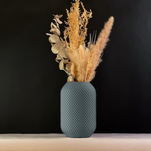 Pode incluir: Um vaso azul-acinzentado texturizado com um padrão de favo de mel, preenchido com gramíneas ornamentais e folhagens secas. O vaso está sobre um fundo escuro. As flores secas são em tons de bege e castanho.