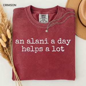 May include: Crimson-colored t-shirt with the phrase "an alani a day helps a lot" in white lettering. The shirt has a crew neck and a small tag at the neckline. A silver necklace with a star pendant is draped over the shirt. A brown hat is visible in the background.