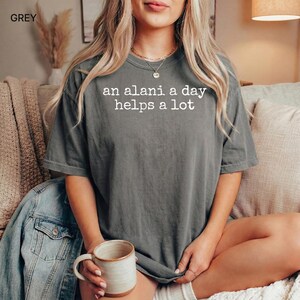 May include: Grey t-shirt with the text "an alani a day helps a lot" in white. The shirt is a relaxed fit and has short sleeves. A ceramic mug with a beige interior is held in the hand. A denim jacket is visible.