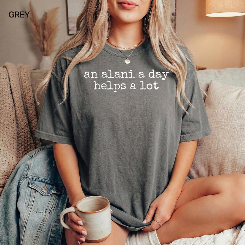 May include: Grey t-shirt with the text "an alani a day helps a lot" in white. The shirt is a relaxed fit and has short sleeves. A ceramic mug with a beige interior is held in the hand. A denim jacket is visible.