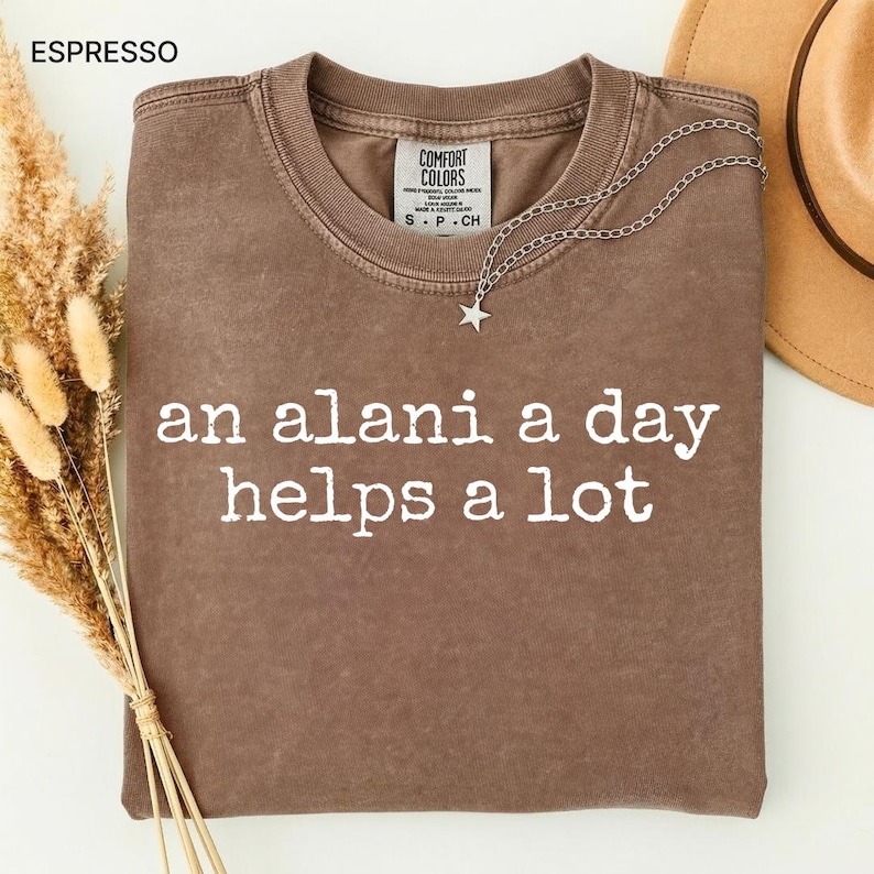 May include: Espresso-colored t-shirt with the text "an alani a day helps a lot" in white. A silver necklace with a star pendant is draped over the shirt. The shirt is folded, and a tan hat is visible in the background.