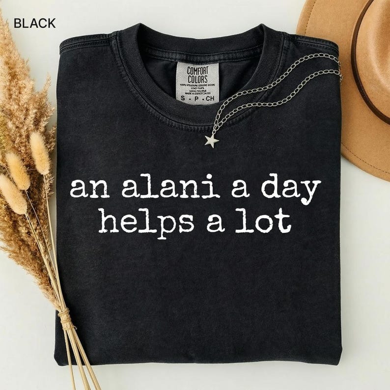 May include: Black Comfort Colors t-shirt with the phrase "an alani a day helps a lot" in white lettering. A silver necklace with a star pendant rests on the shirt. A beige hat and dried flowers are in the background.