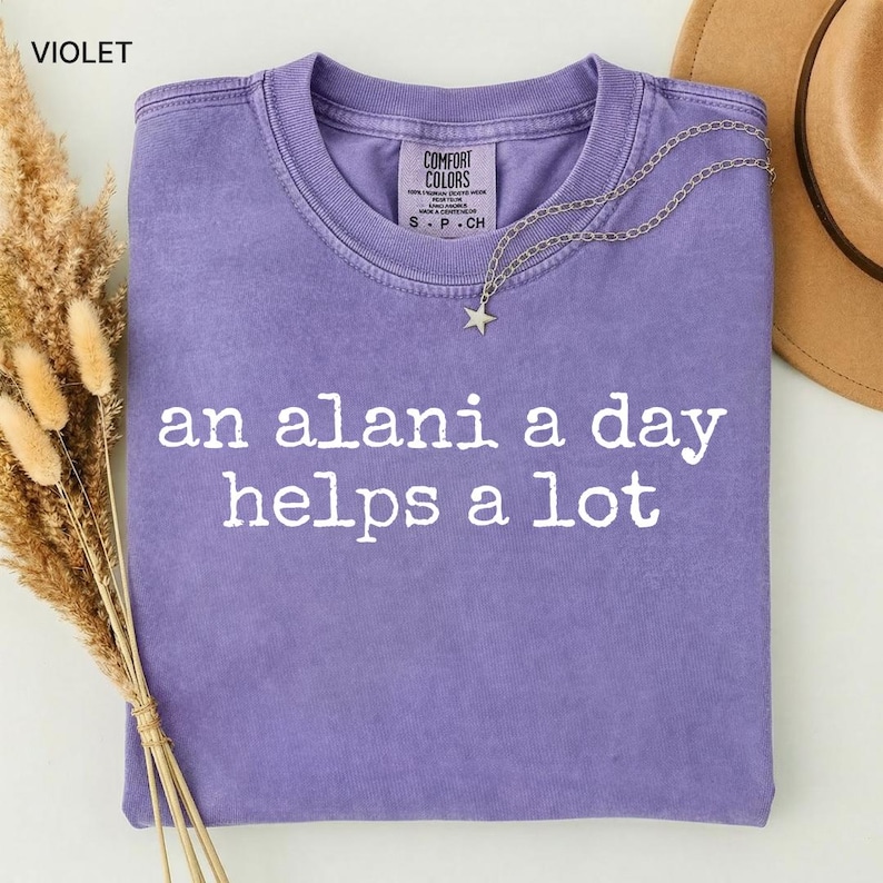 May include: Violet-colored t-shirt with the text "an alani a day helps a lot" in white. A silver necklace with a star pendant rests on the shirt. The shirt is folded, and a brown hat is visible in the background.