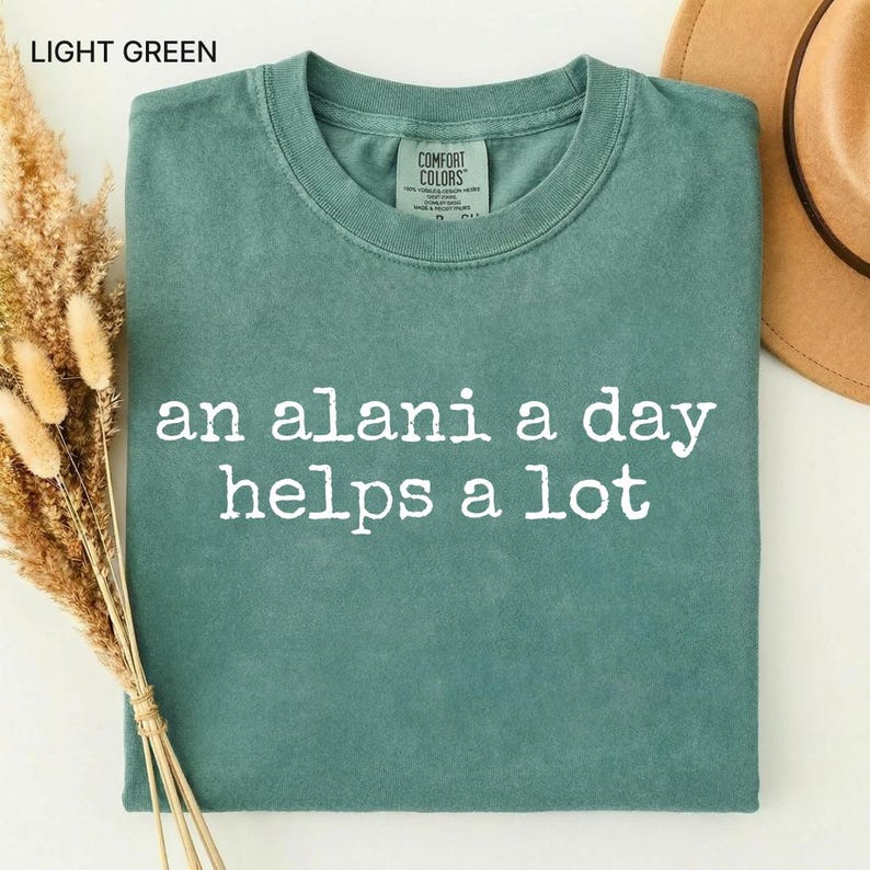 May include: Light green t-shirt with the text "an alani a day helps a lot" in white. The shirt is folded and has a Comfort Colors tag. A brown hat and dried flowers are in the background.