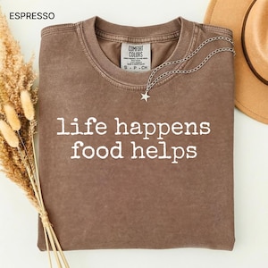 以下が含まれることがあります： 「life happens food helps」の白い文字がプリントされたエスプレッソ色のTシャツ。星のペンダントが付いたシルバーのネックレスがシャツの上にかけられています。背景にはベージュの帽子が見えます。