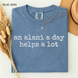 May include: Blue jean colored Comfort Colors t-shirt with the text "an alani a day helps a lot" in white. A silver necklace with a star pendant and a brown hat are on the shirt. Dried flowers are on the left side.