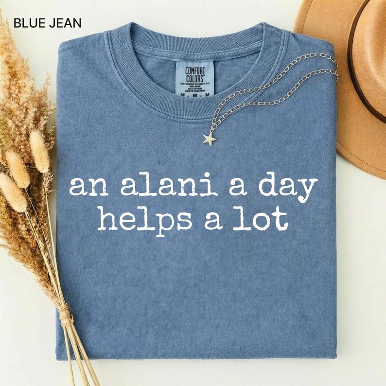 May include: Blue jean colored Comfort Colors t-shirt with the text "an alani a day helps a lot" in white. A silver necklace with a star pendant and a brown hat are on the shirt. Dried flowers are on the left side.