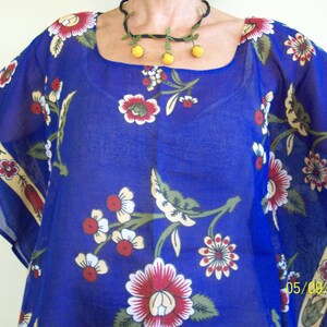 May include: A blue sheer top with a floral print and a yellow beaded necklace. The top has a square neckline and short sleeves. The floral print features red, white, and green flowers.