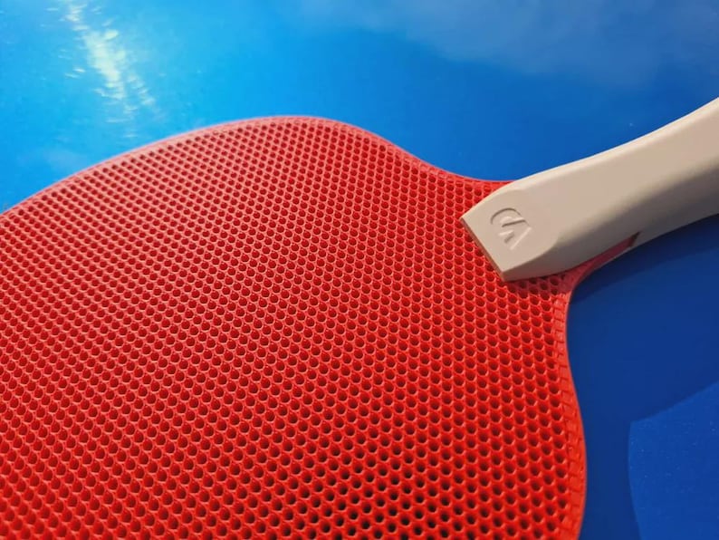 May include: Close-up of a red ping pong paddle with a perforated surface and a gray handle. The handle has a white logo. The paddle is set against a blue background.