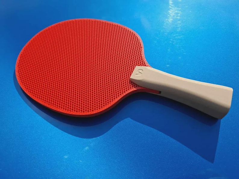 May include: A red ping pong paddle with a gray handle. The paddle has a textured surface and is set against a blue background. The handle has a curved design.