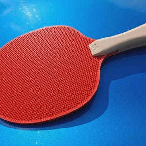 May include: A red ping pong paddle with a textured surface and a light gray handle. The paddle is set against a blue background, with a shadow cast to the side. The handle has a small logo.